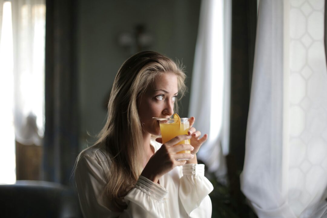 Current image: A thoughtful woman drinks orange juice by a window, embodying calm and refreshment indoors.